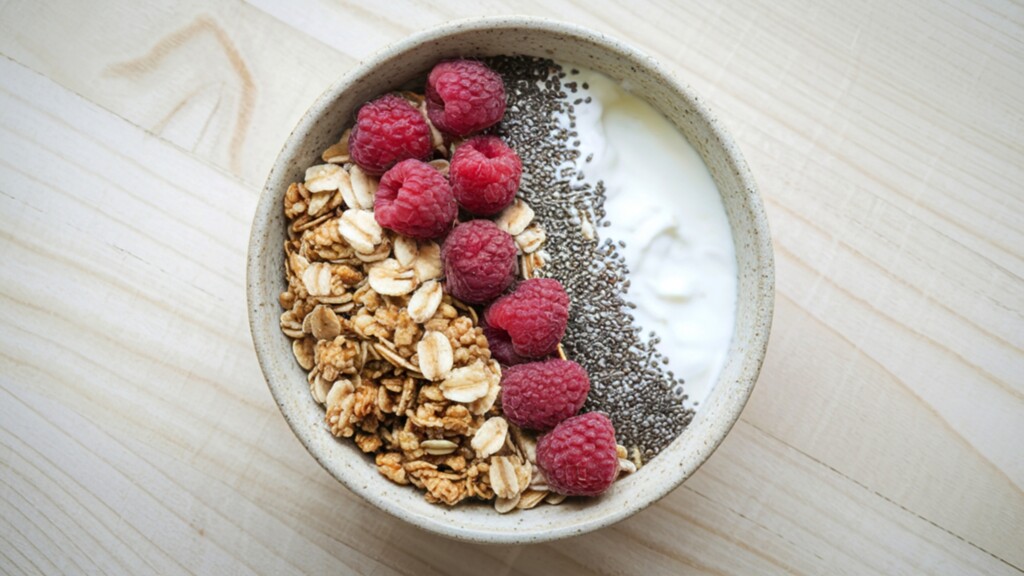 Yogurt bowl topped with granola, raspberries, oats, and chia seeds on a light wooden table.