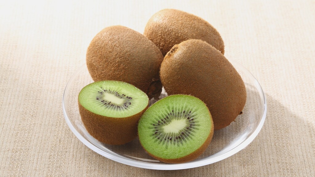 Fresh kiwi halves and whole kiwis in a glass bowl on a neutral linen surface.