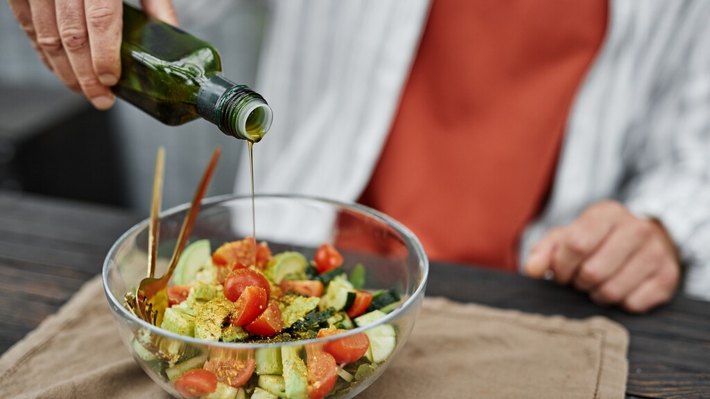 Olive oil being poured over a fresh Mediterranean salad with tomatoes, cucumber, avocado, herbs, and spices.