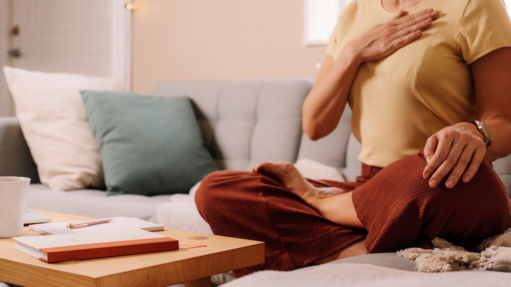 Woman practicing mindful breathing on a living room sofa with one hand on her chest, using a calm posture to support relaxation and digestion.
