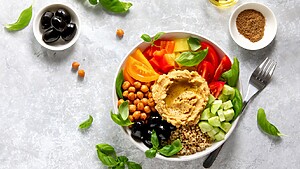 Mediterranean quinoa bowl with hummus, chickpeas, tomatoes, cucumber, black olives, basil, and olive oil on a gray table.