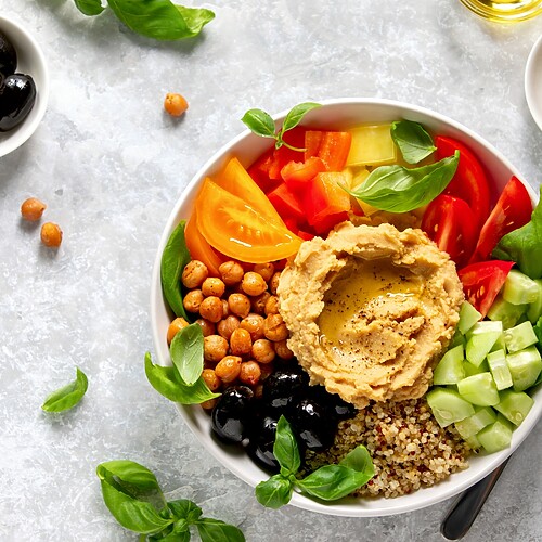 Mediterranean quinoa bowl with hummus, chickpeas, tomatoes, cucumber, black olives, basil, and olive oil on a gray table.