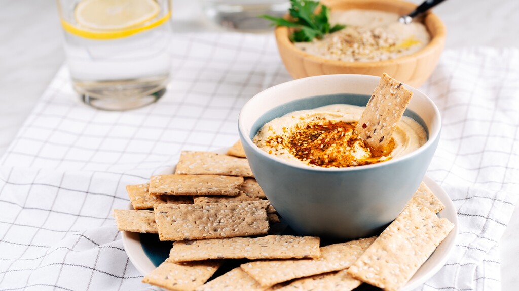 Bowl of creamy hummus topped with spices and olive oil, served with seeded crackers on a light tablecloth.
