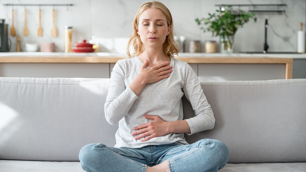 Woman sitting cross-legged on a sofa with eyes closed, placing one hand on her chest and one on her stomach while practicing calming breathwork.