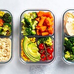 Three glass meal prep containers filled with chicken, avocado, broccoli, tomatoes, and prepared vegetables.