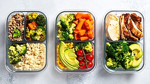 Three glass meal prep containers filled with chicken, avocado, broccoli, tomatoes, and prepared vegetables.