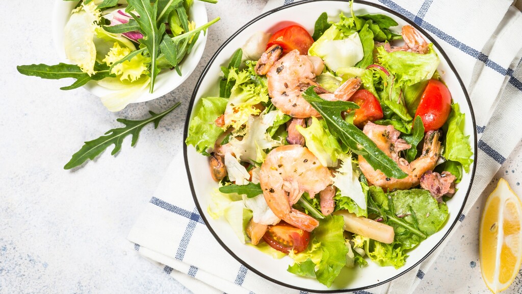 Shrimp salad with leafy greens, tomatoes, and lemon wedges in a ceramic bowl.