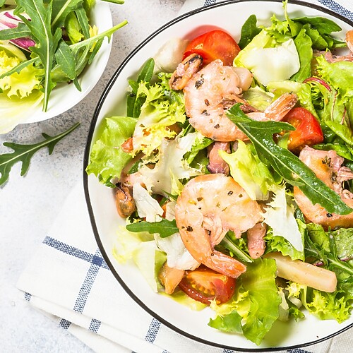 Shrimp salad with leafy greens, tomatoes, and lemon wedges in a ceramic bowl.