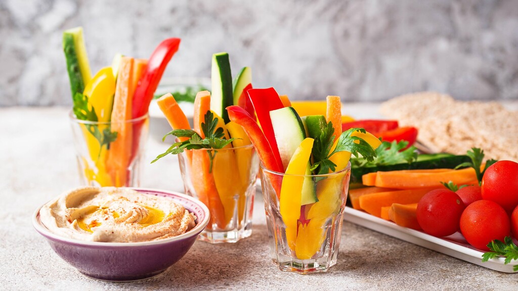 Colorful vegetable sticks with hummus, whole grain crackers, cherry tomatoes, and fresh herbs on a light stone surface.