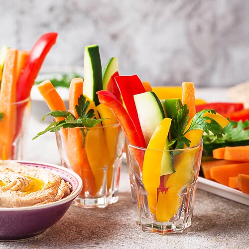 Colorful vegetable sticks with hummus, whole grain crackers, cherry tomatoes, and fresh herbs on a light stone surface.