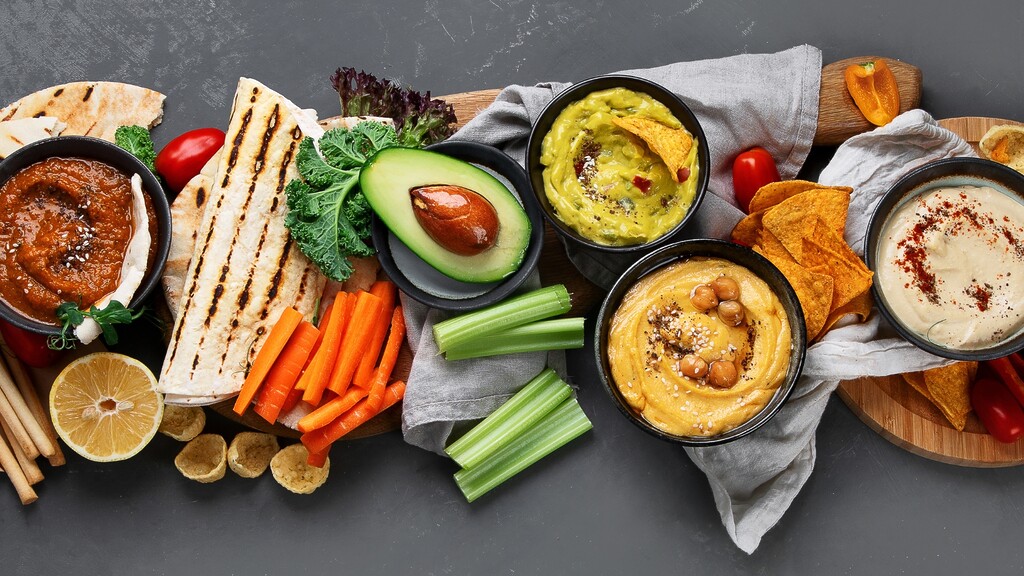 Mediterranean snack spread with hummus, vegetable dips, grilled pita, avocado, chips, carrots, celery, tomatoes, and lemon.