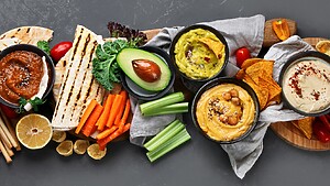 Mediterranean snack spread with hummus, vegetable dips, grilled pita, avocado, chips, carrots, celery, tomatoes, and lemon.