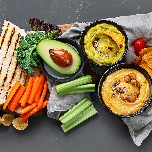 Mediterranean snack spread with hummus, vegetable dips, grilled pita, avocado, chips, carrots, celery, tomatoes, and lemon.