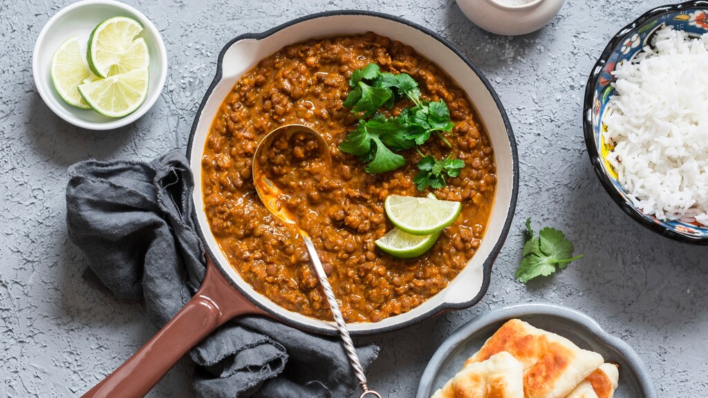 Lentil stew served in a pan with lime wedges, cilantro, rice, and flatbread on a gray tabletop.