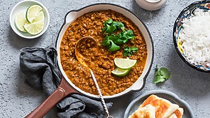 Lentil stew served in a pan with lime wedges, cilantro, rice, and flatbread on a gray tabletop.