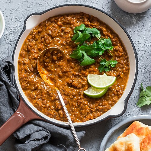 Lentil stew served in a pan with lime wedges, cilantro, rice, and flatbread on a gray tabletop.