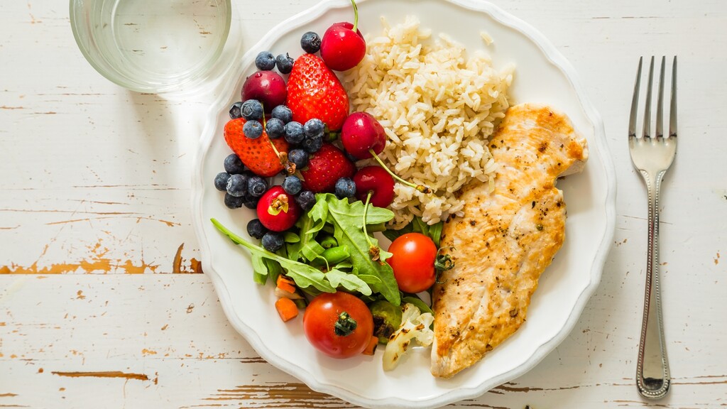Healthy balanced plate with grilled chicken, quinoa, avocado, sprouts, arugula, tomatoes, and lime on a blue textured table.