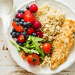 Healthy balanced plate with grilled chicken, quinoa, avocado, sprouts, arugula, tomatoes, and lime on a blue textured table.