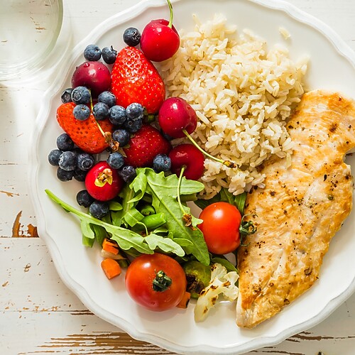 Healthy balanced plate with grilled chicken, quinoa, avocado, sprouts, arugula, tomatoes, and lime on a blue textured table.