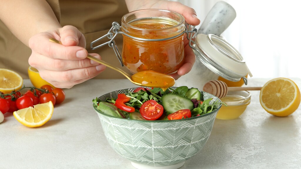 Homemade vinaigrette being spooned over a fresh salad with cucumbers, tomatoes, lemon, and honey.