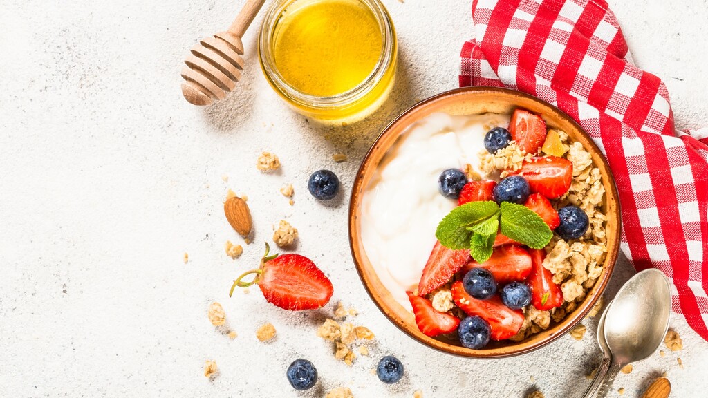 Plain Greek yogurt bowl topped with strawberries, blueberries, granola, mint, and honey on a light breakfast table.