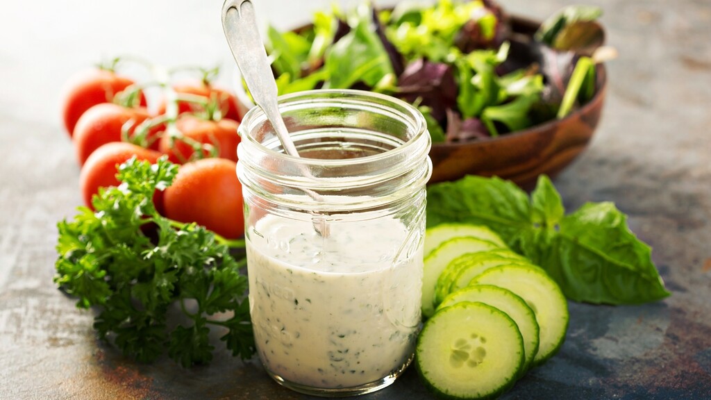 Creamy herb dressing in a glass jar with cucumber slices, fresh herbs, tomatoes, and salad greens. 