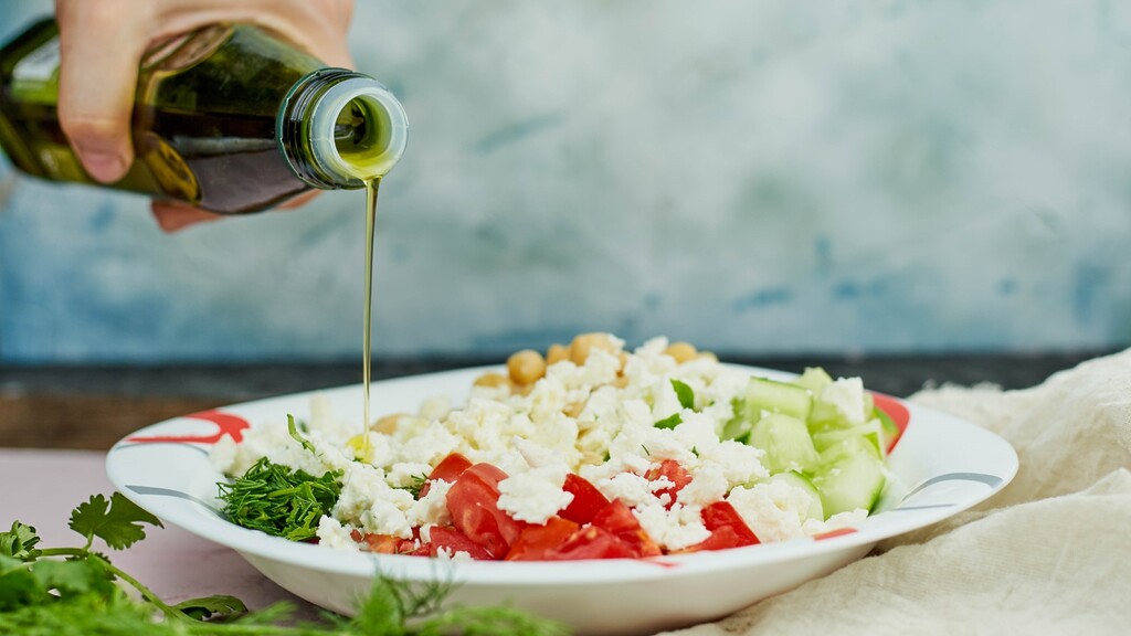Extra virgin olive oil being poured over a Mediterranean salad with chickpeas, feta, tomatoes, and cucumber.