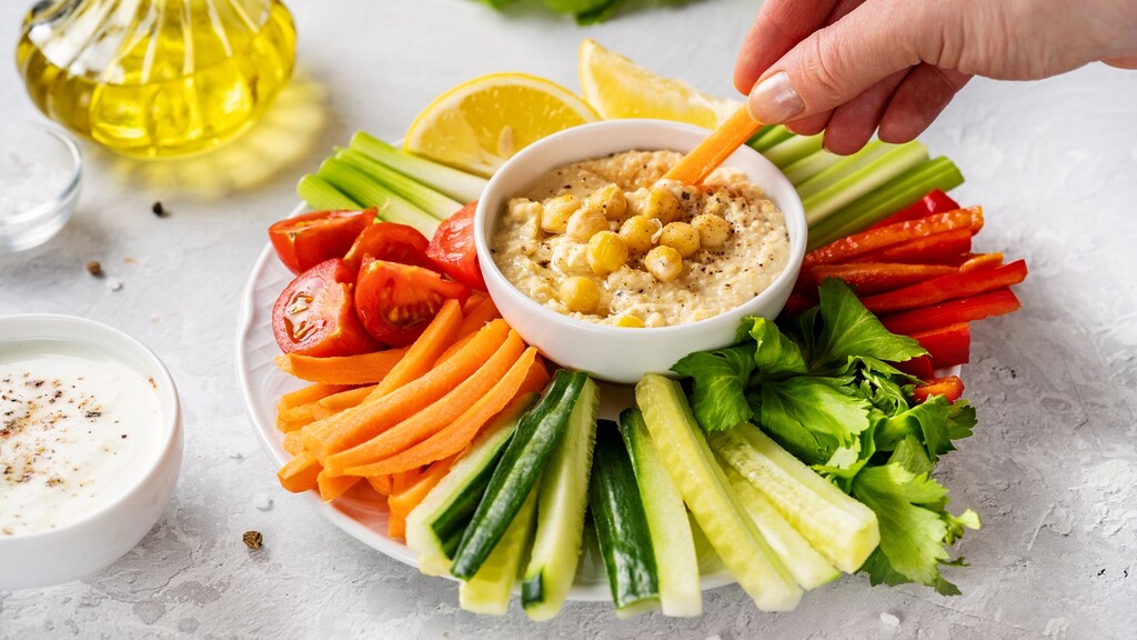 Carrot stick dipped into chickpea hummus with cucumber, celery, bell pepper, tomatoes, lemon, and olive oil.