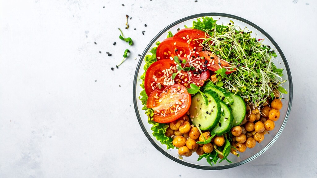 Chickpea salad with sliced tomatoes, cucumber, greens, sprouts, and sesame seeds in a glass bowl.