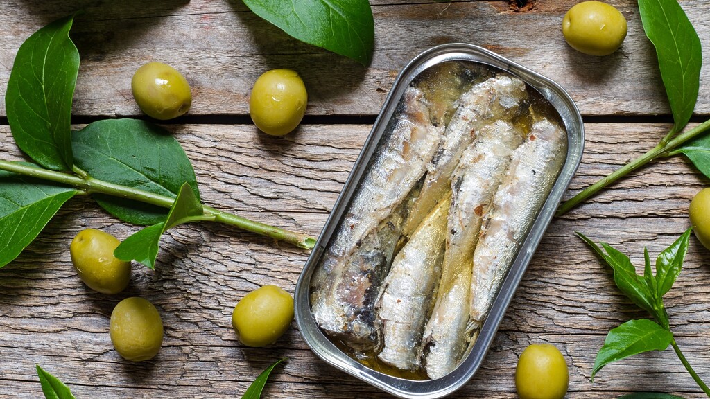 Open tin of sardines with green olives and herbs on a rustic wooden table.