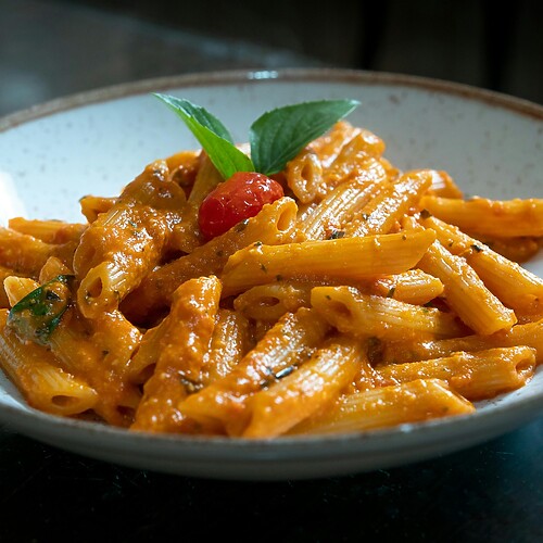 Penne pasta in creamy tomato sauce served in a white bowl with basil and a cherry tomato garnish.