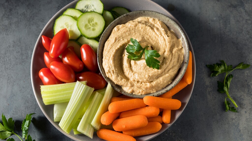 Hummus bowl with cucumber slices, cherry tomatoes, celery sticks, and baby carrots on a gray plate.