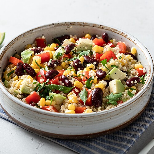 Quinoa salad with kidney beans, corn, avocado, red peppers, herbs, and lime wedges in a ceramic bowl.