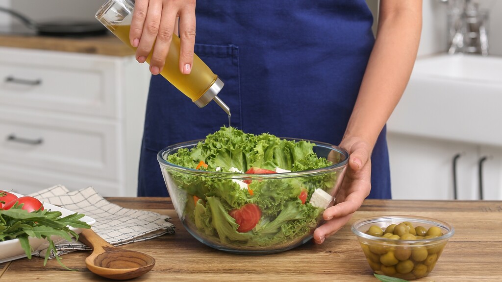 Olive oil being poured over a fresh green salad with tomatoes, feta, and olives in a kitchen.