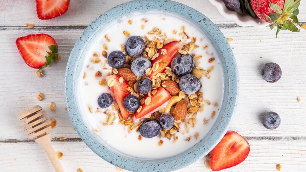 Greek yogurt bowl topped with blueberries, strawberries, almonds, granola, and honey on a white wooden table.