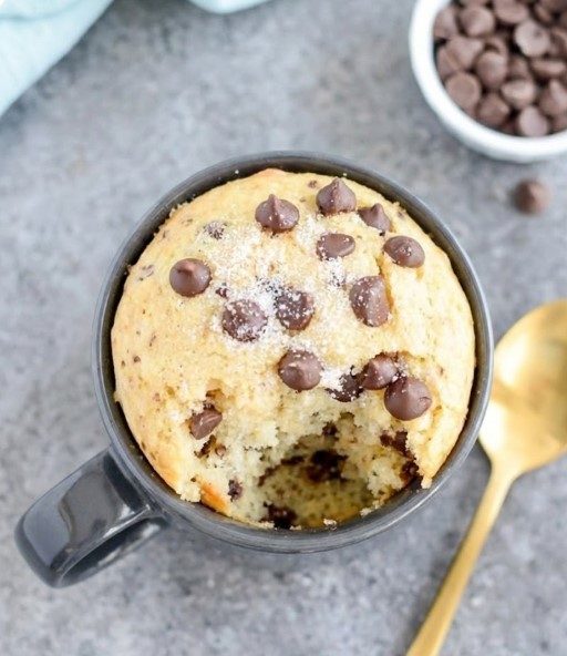 Low Carb Chocolate Chip Mug Cake - a warm, fluffy vanilla cake served in a dark grey ceramic mug, generously topped with sugar-free chocolate chips and a sprinkle of granular sweetener, with a large bite revealing the tender, tender crumb inside