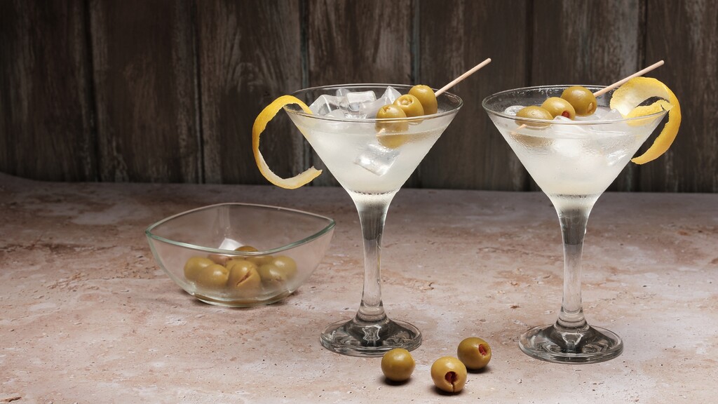 Two dirty martinis with olives and lemon twists on a rustic bar table.