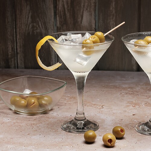 Two dirty martinis with olives and lemon twists on a rustic bar table.