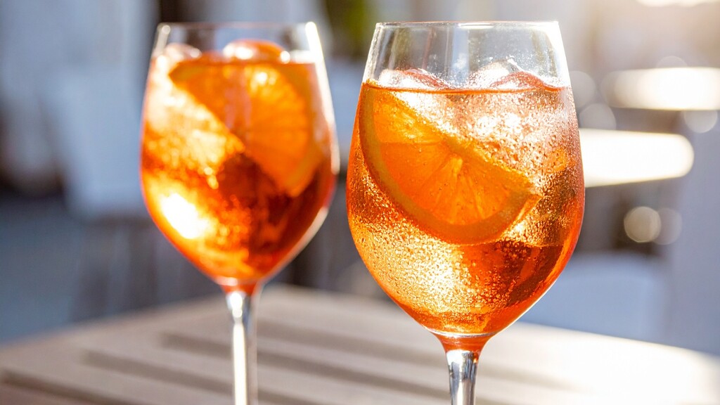 Two Aperol spritz cocktails with orange slices and ice in sunlit wine glasses.