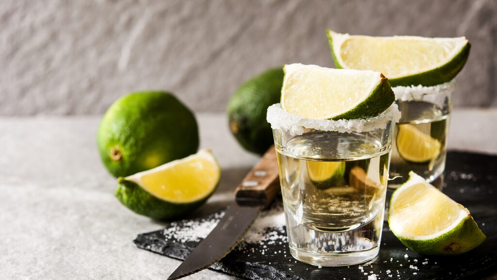 Blanco tequila shots with lime wedges and salt on a stone surface.
