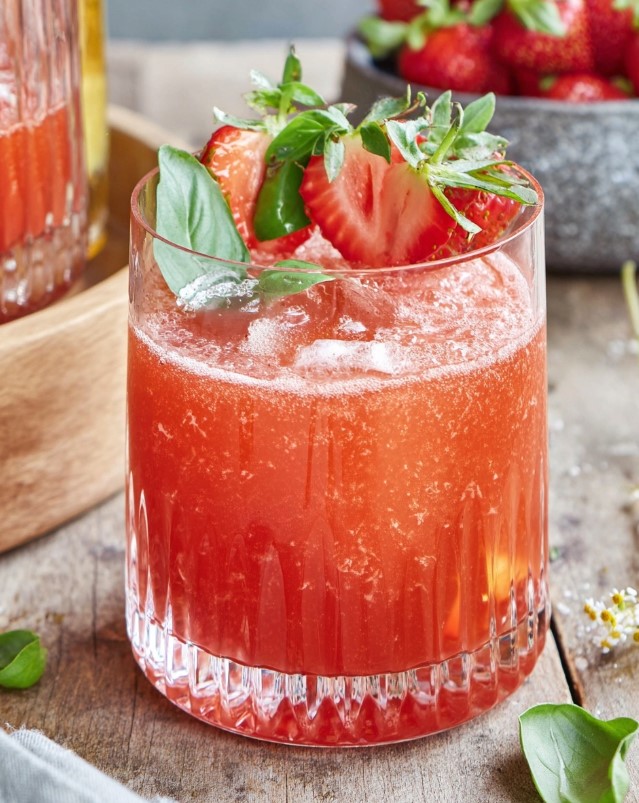 Pink iced cocktail in a low glass garnished with strawberry slices and fresh basil leaves