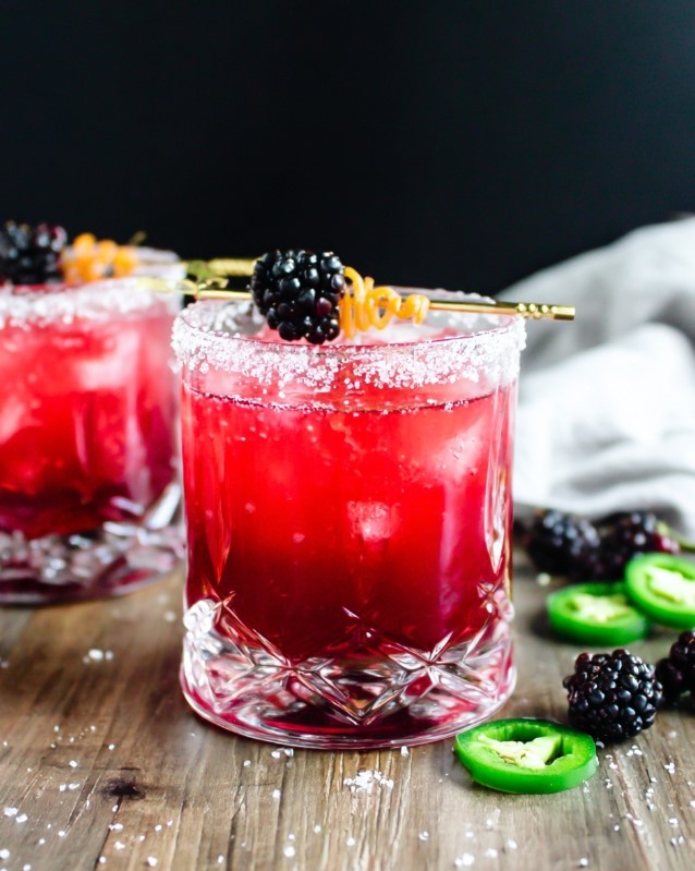 Spicy blackberry margarita cocktail ready to serve in a rocks glass with a salted rim, ice, garnished with a blackberry and orange peel twist, with jalapeño slices on the side